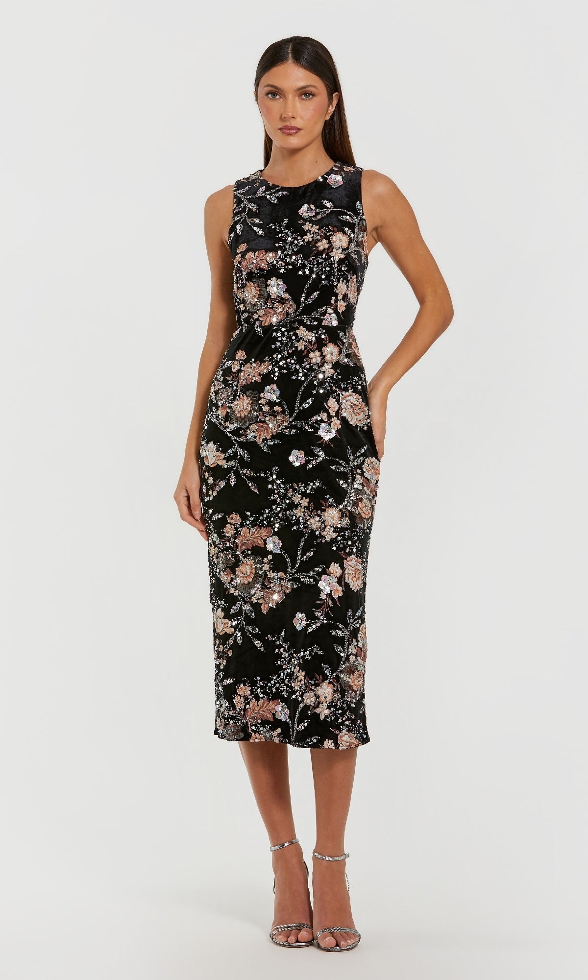 A woman stands against a plain white background wearing the Mac Duggal 68958, a sleeveless black midi wedding guest dress with a floral pattern, paired with silver high-heeled sandals.