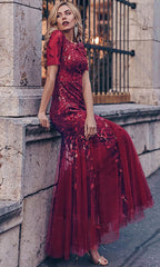 A woman in a Short-Sleeve Modest Long Mermaid Prom Dress 7707 with sheer embroidered floral details sits on a stone ledge in front of a wrought iron fence.