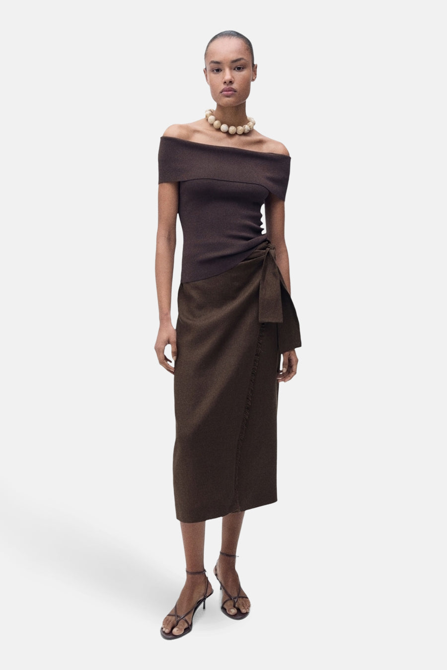 Bruine rok van MANGO, met knoopdetail aan de zijkant, gecombineerd met off-shoulder top en haksandalen.
