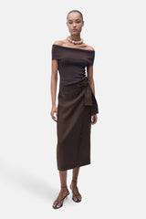 Bruine rok van MANGO, met knoopdetail aan de zijkant, gecombineerd met off-shoulder top en haksandalen.
