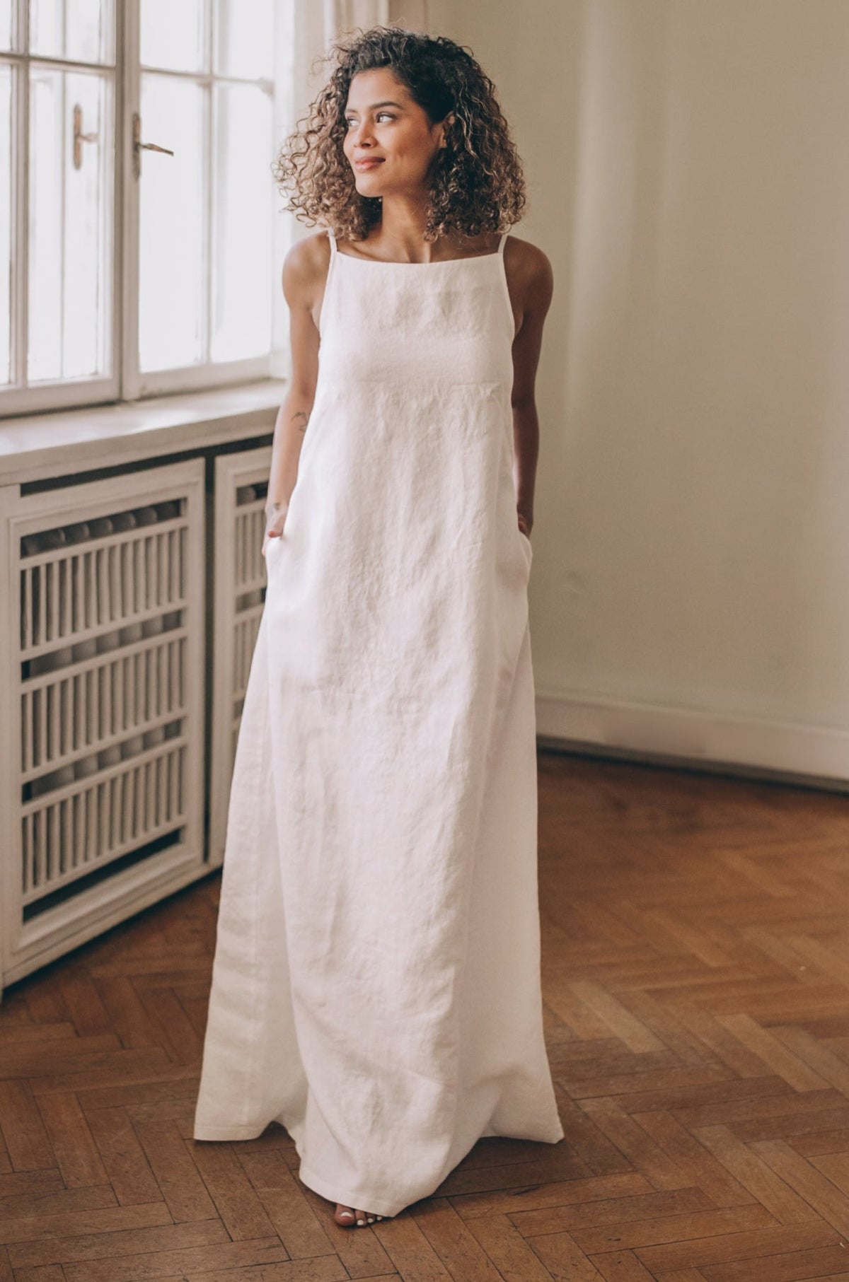 Woman wearing sleeveless linen maxi dress with boat neckline and pockets standing indoors
