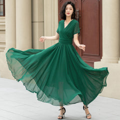 A woman wearing a deep green chiffon V-neck maxi dress with short sleeves and an ankle-length silhouette, standing in front of a building.