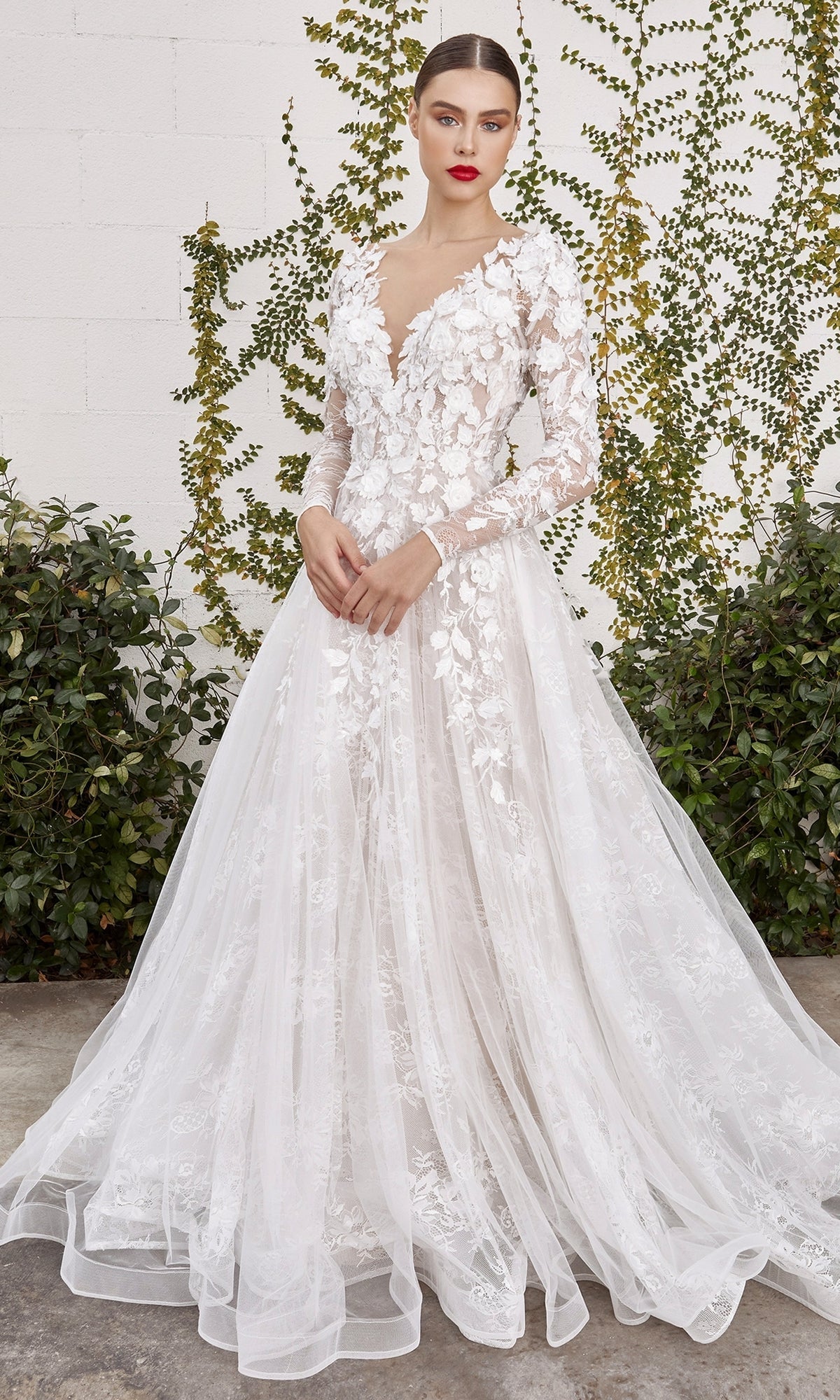Someone stands outside in front of a greenery-covered white brick wall, wearing the LadivineA1067W Long Sleeve White Lace Bridal Gown, which features floral patterns and a deep V-neckline.