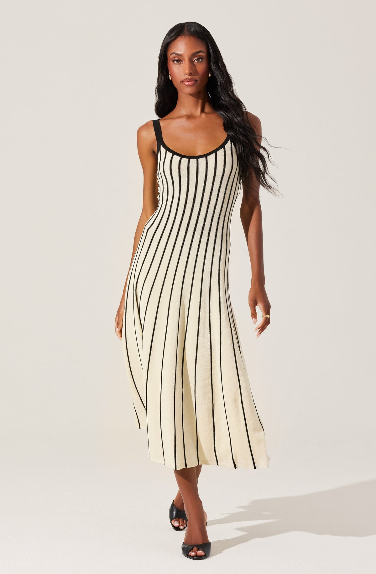 A woman is wearing a sleeveless, ankle-length dress with vertical black and beige stripes. The dress has black shoulder straps and black trim around the neckline and armholes. She is also wearing black high-heeled sandals.