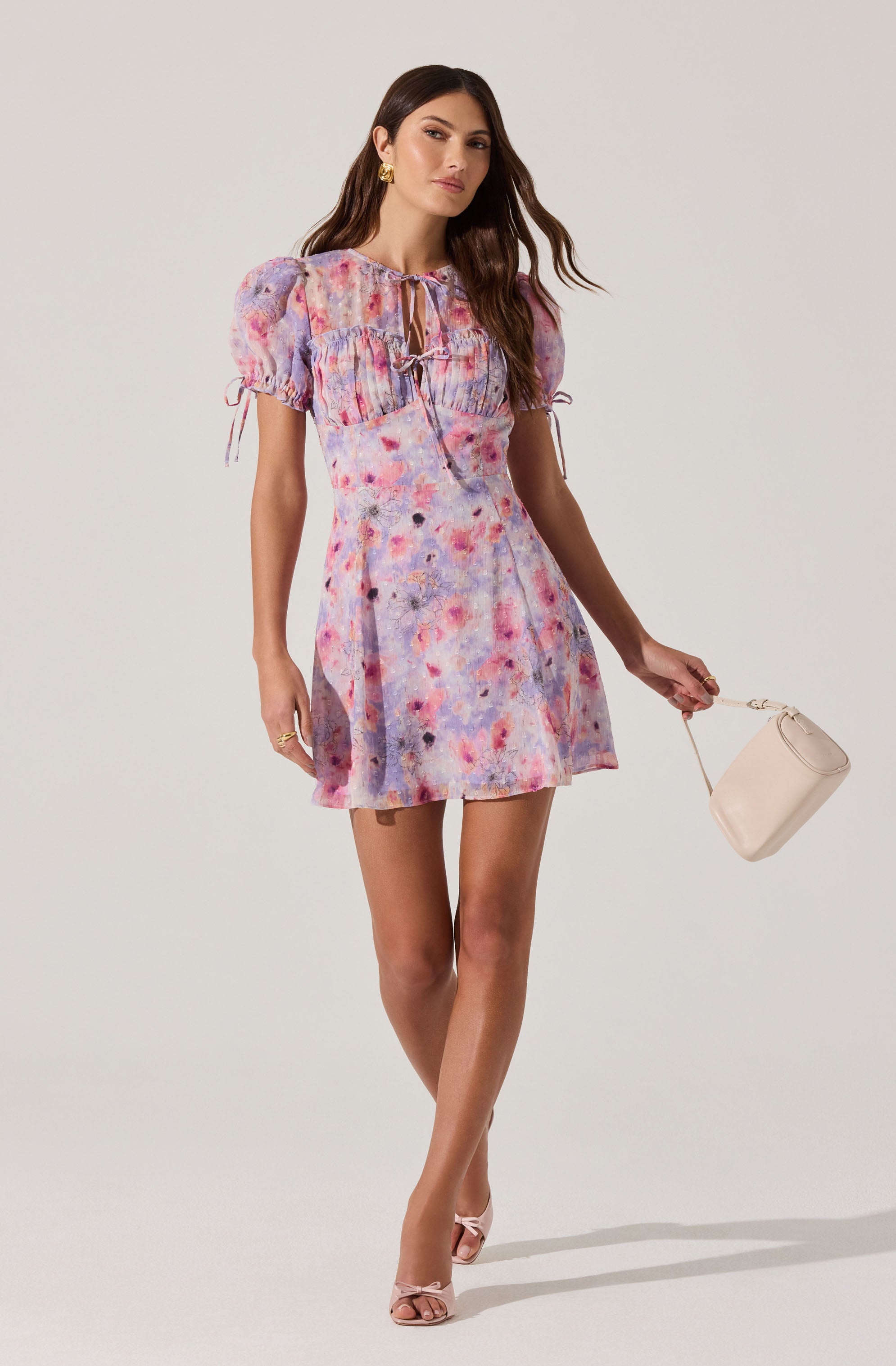 A woman is wearing a colorful, watercolor-like patterned dress with shades of pink, purple, and blue. The dress has puffed short sleeves with adjustable drawstrings, a fitted waist, and a short, above-the-knee hemline. She is accessorizing with gold earrings and rings, and holding a small beige handbag. She is also wearing light pink open-toe heels.