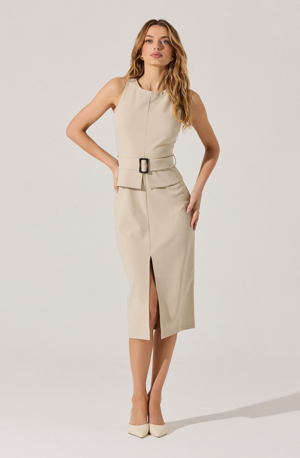 A woman is wearing a sleeveless beige dress with a fitted waist and a wide belt with a rectangular buckle. The dress features a front slit that extends below the knees. She is also wearing matching beige pointed-toe heels.
