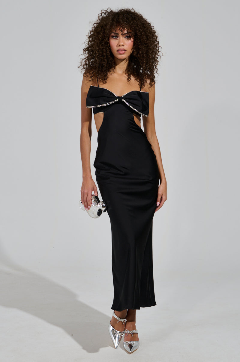 A person with curly hair is wearing the "Put a Bow on It Satin Maxi Dress in Black," featuring cut-out details and rhinestone accents, ideal for a holiday party. They are holding a shiny silver clutch and wearing pointed silver shoes against a plain background.