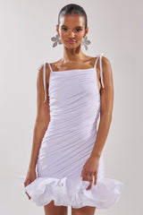 A woman wears the PRINCESS PEPLUM MINI DRESS IN WHITE, featuring a fitted ruched mesh design with shoulder tie straps and an organza ruffle hemline. Her hair is slicked back and she accessorizes with large floral statement earrings.