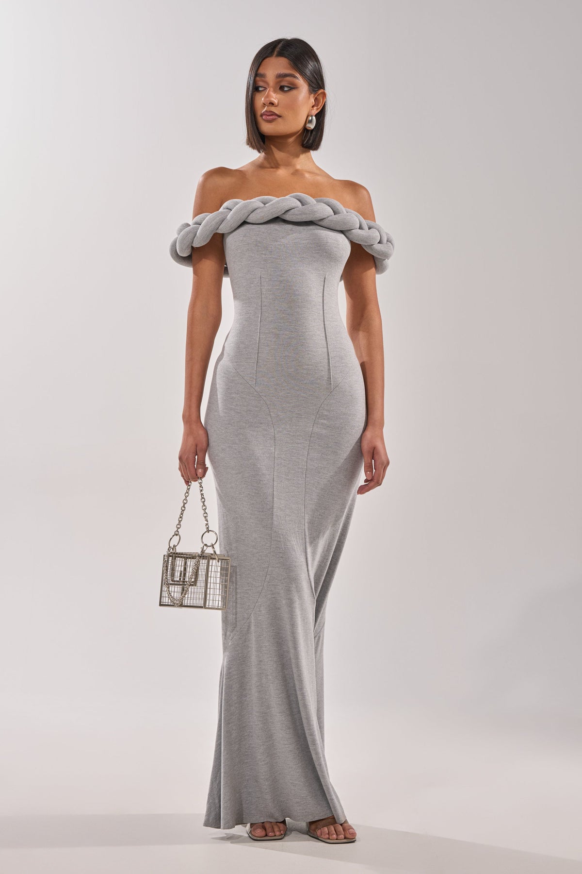 A woman in the GODDESS HOUR KNIT MAXI DRESS stands holding a metallic box purse and wears strappy heels. With straight dark hair, statement earrings, and an off-the-shoulder style, she radiates modern elegance.