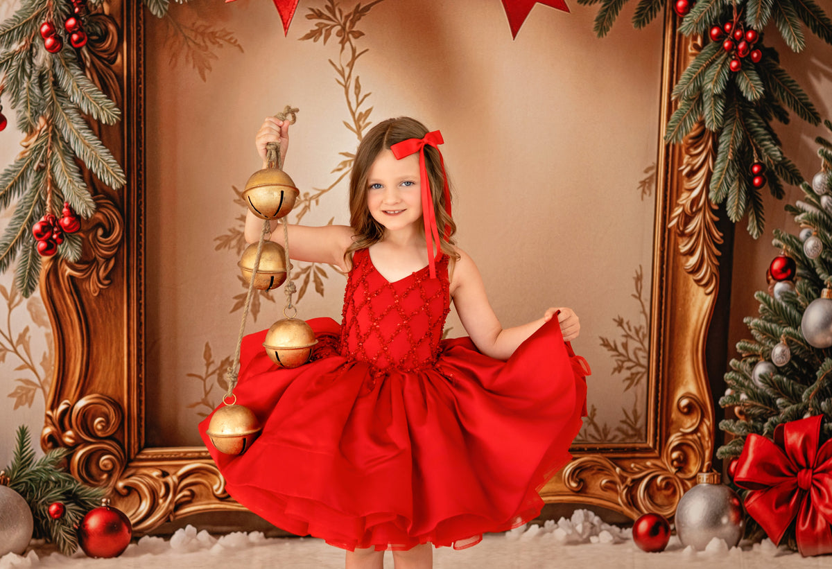 Young girl in a red dress holding Christmas ornaments in a decorated setting with a gold frame and festive decorations.