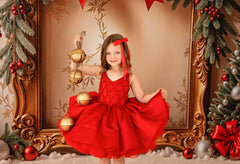 Young girl in a red dress holding Christmas ornaments in a decorated setting with a gold frame and festive decorations.