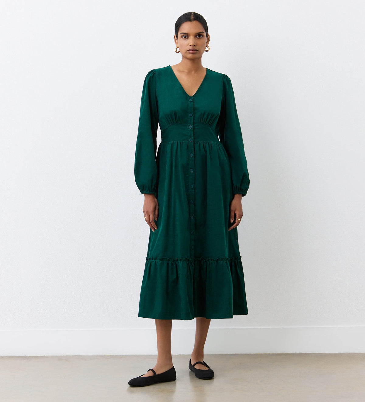 A woman stands against a white wall wearing the Arizona Bottle Green Corduroy Midi Dress, featuring long sleeves, an empire waistband, and ruffled hem, paired with black flats and gold hoop earrings.