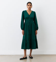 A woman stands against a white wall wearing the Arizona Bottle Green Corduroy Midi Dress, featuring long sleeves, an empire waistband, and ruffled hem, paired with black flats and gold hoop earrings.