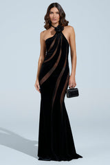 Against a light blue backdrop, a woman poses in the SEE THROUGH ME MAXI DRESS, featuring floor-length black velvet and sheer diagonal stripes. She carries a small black handbag and has wavy brown hair, exuding elegance with the gown's mermaid silhouette.