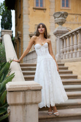 A woman wearing a white bridal dress with a sweetheart neckline and lace details, standing on steps with a garden and old building in the background.