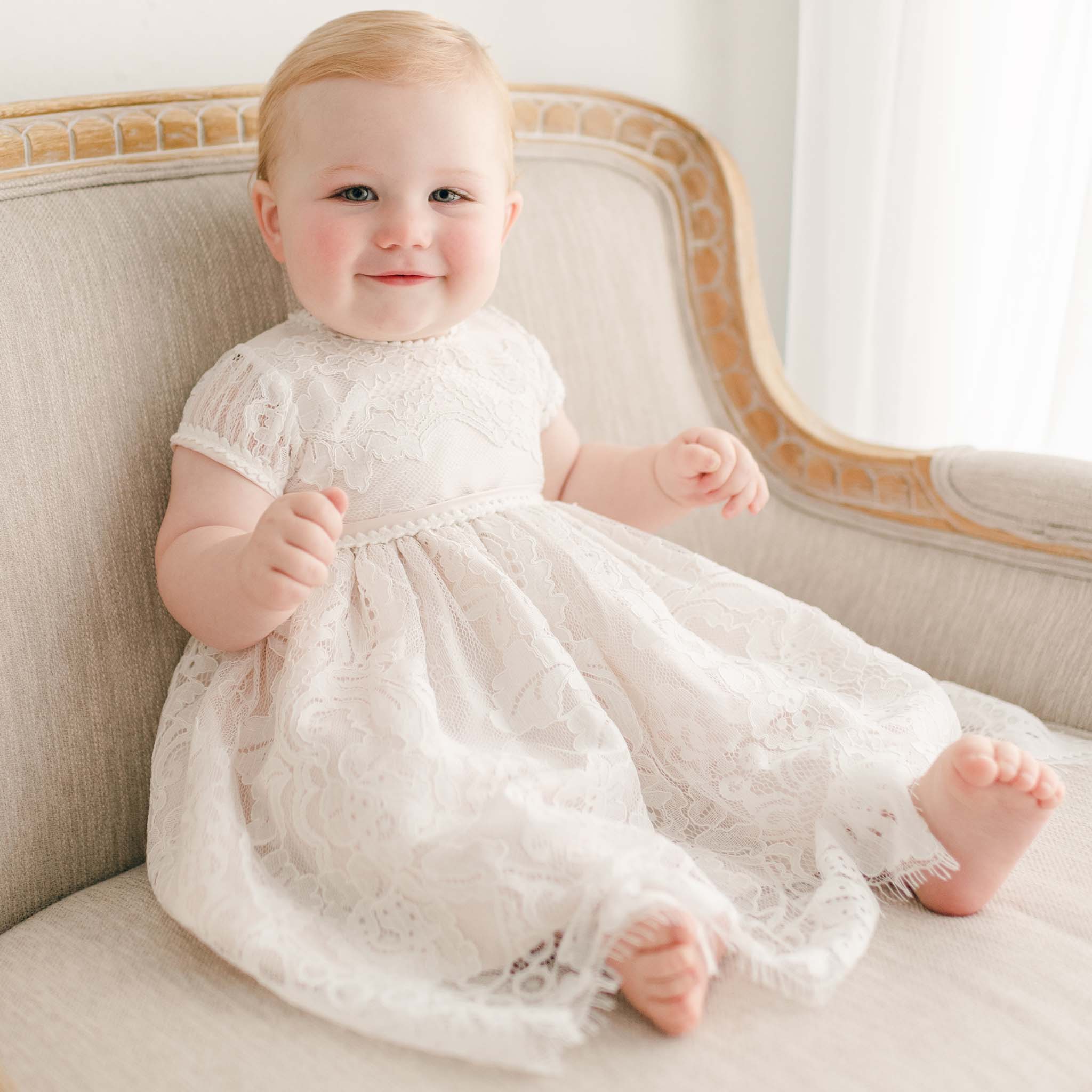 A cheerful baby with light blonde hair sits on a beige fabric sofa, wearing the Victoria Puff Sleeve Dress adorned with embroidered lace, and looks toward the camera. The softly lit setting creates a warm ambiance in the scene.