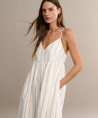 A woman with long brown hair wears the Cove Dress, a sleeveless casual dress with slender straps and thin gold stripes, standing with one hand in her pocket against a beige background.