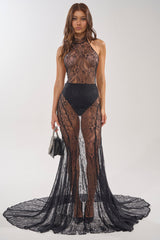 A woman faces forward in the NEVER BETTER MOCK NECK LACE MAXI DRESS IN BLACK, featuring a high neckline and flowing train. She holds a small black handbag, her long wavy hair down against a plain light background.
