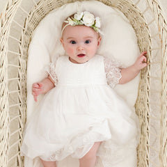 A baby wearing a vintage-inspired Ella baptism dress and flower headband lies in a wicker bassinet.