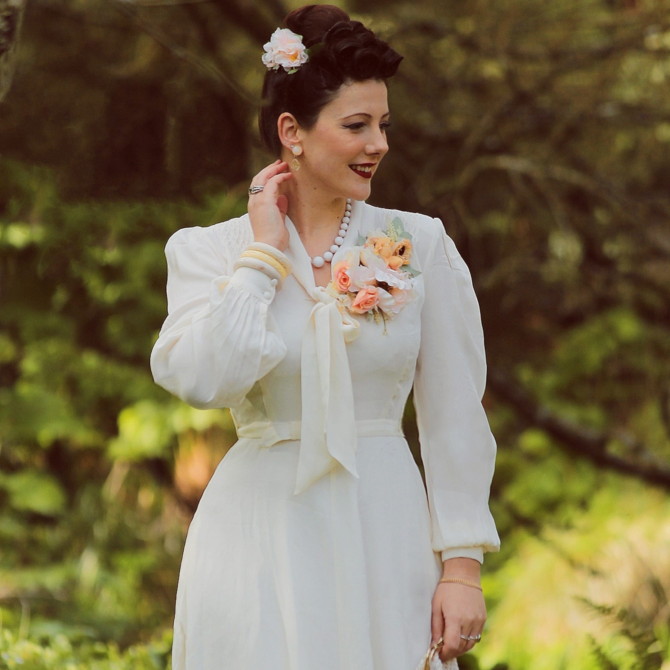 A woman wearing a vintage-inspired cream dress with a pussy bow neckline, long sleeves, and smock stitching on the shoulders. The dress features button fastenings, a side zip, waist belt, and side-seam pockets, paired with a complimentary slip.