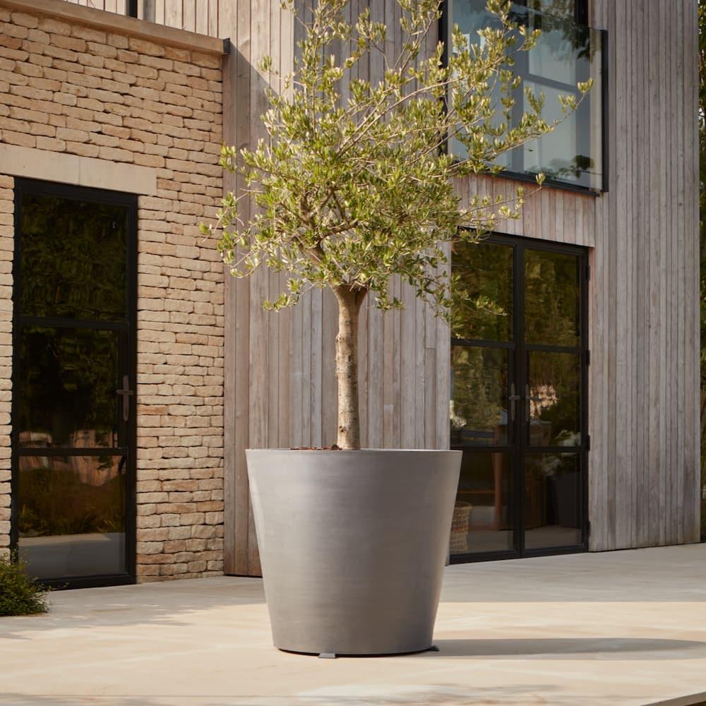 Tall contemporary plastic planter on wheels with a sleek matte grey finish, styled with a tree on a modern patio for a minimalist outdoor aesthetic.