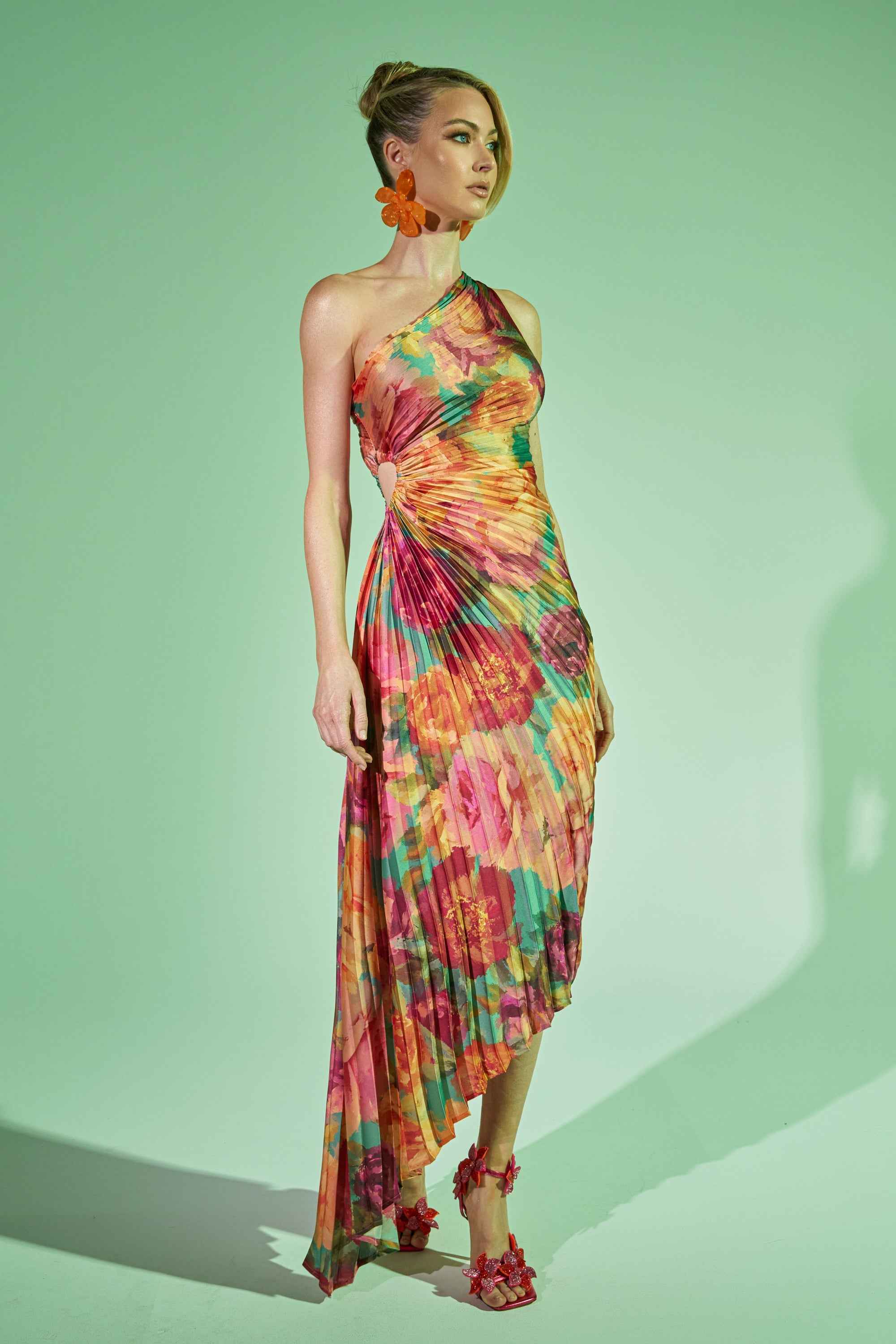A woman stands against a green background in the GARDEN FLORAL PLEATED MIDI DRESS, featuring a one-shoulder design, pleats, an abstract floral print, and an asymmetrical hem. She pairs it with matching earrings and heels; her hair is styled up.
