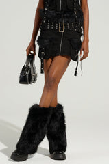 A person in the DELANEY FUR TRIM MINI SKIRT with metal eyelets, styled with matching black fur boots and a small black handbag, standing against a plain, light-colored background.