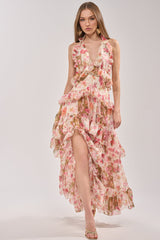 A woman wears the SUMMER STROLL FLORAL CHIFFON MAXI DRESS, featuring pink floral chiffon, ruffled layers, a deep V-neck, and a high slit. She stands against a light background in sandals—ideal for special occasions.