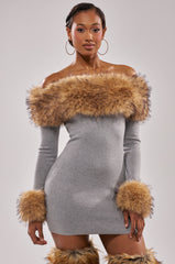 A woman models the COMFY COZY FUR TRIM MINI DRESS IN GREY, featuring a fitted ribbed knit, off-shoulder cut, and fur trim on the neckline and cuffs, paired with matching boots against a light background.