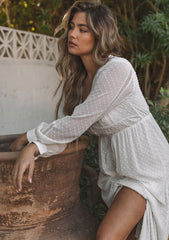 [Color: Gardenia] A model wearing a dreamy sheer off white maxi dress in swiss dot chiffon. With lace trim details throughout, a self covered button front, a sexy front slit, and sheer voluminous long sleeves with a button wrist cuff.