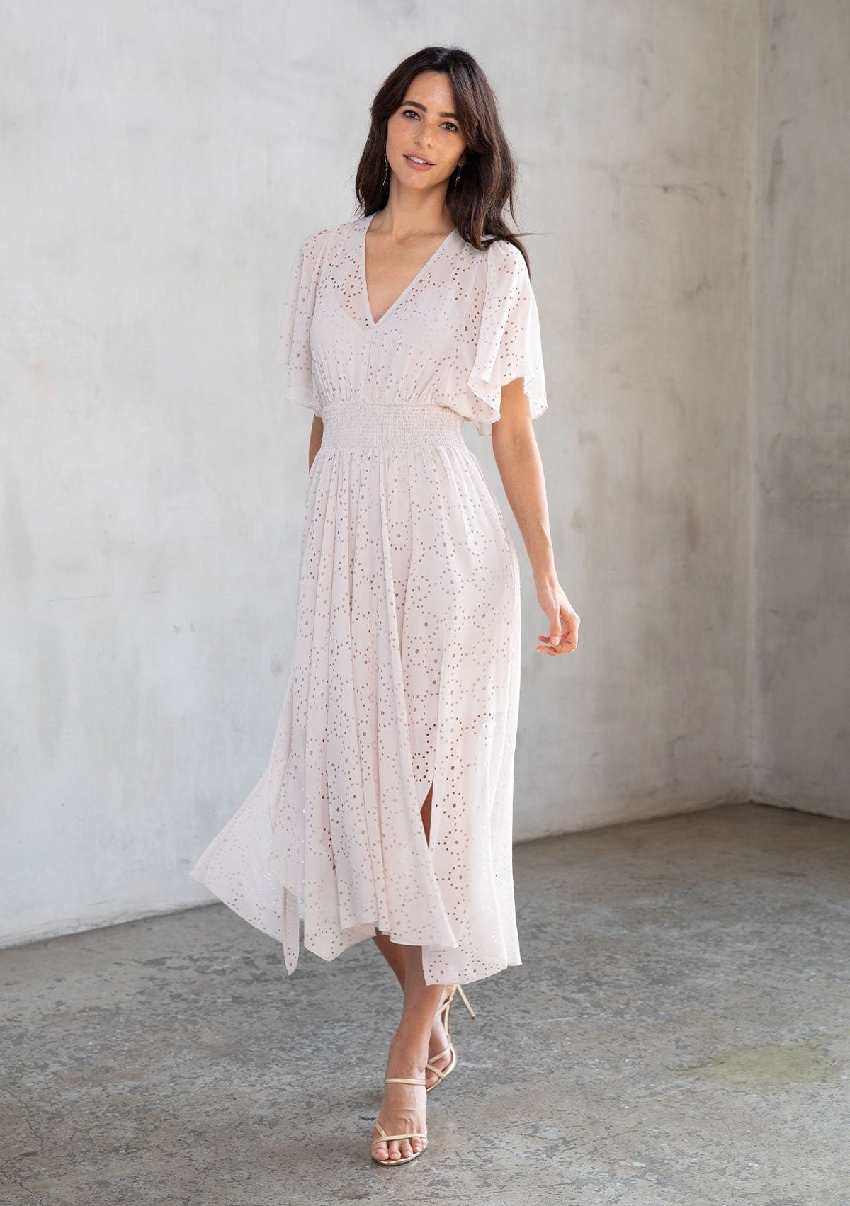 [Color: Bone] A model wearing a dreamy sheer off white chiffon maxi dress with gold trimmed laser cut details throughout. With a flowy skirt, short flutter sleeves, and a shark bite hemline.