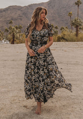 [Color: Black/Lemon] A model wearing a dreamy flowy bohemian maxi dress in a black and yellow floral print. With short puff sleeves, sheer lace accents, and a button front.