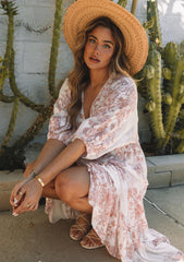 [Color: Ivory/Light Rust] A front facing image of a blonde model sitting outside wearing a bohemian spring maxi dress in an ivory and light pink floral print. With half length puff sleeves, a surplice v neckline, a flowy tiered skirt, and a drawstring waistline with side tie details. Featuring little pom trim throughout.
