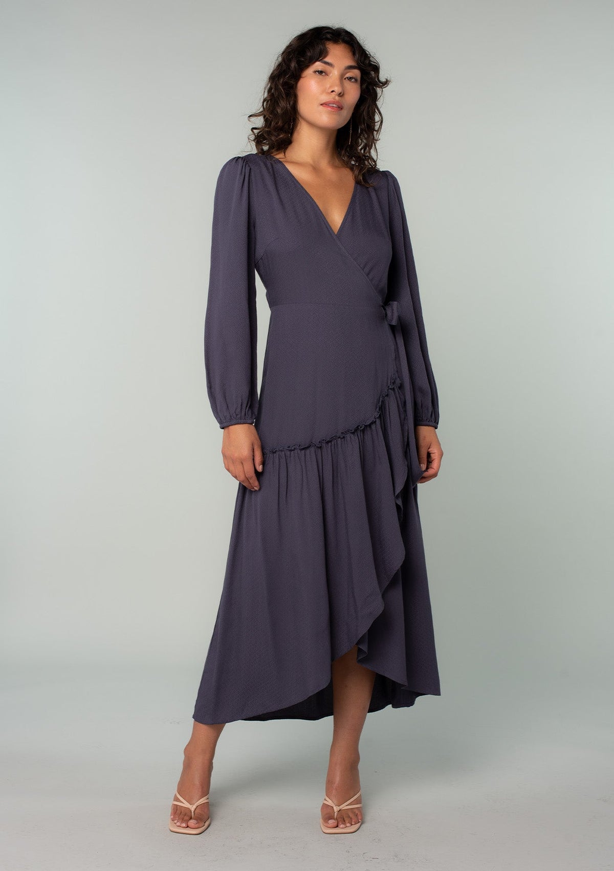 [Color: Periscope Grey] A front facing image of a brunette model wearing a dark grey maxi wrap dress with long sleeves, a ruffle trimmed tiered high low hemline, and a side tie closure.