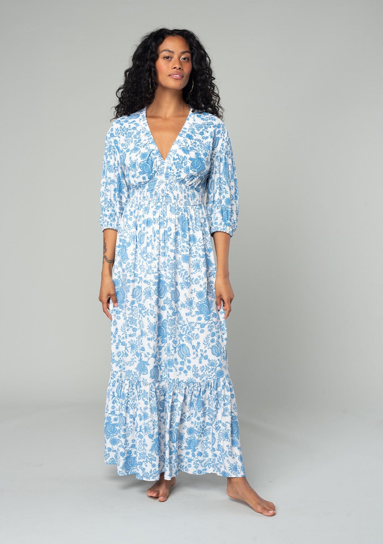 [Color: Cream/Dusty Blue] A front facing image of a brunette model wearing a bohemian cottage core style maxi dress in a white and blue floral print. With half length puff sleeves, a smocked elastic waist detail, a flowy tiered skirt, and a v neckline.
