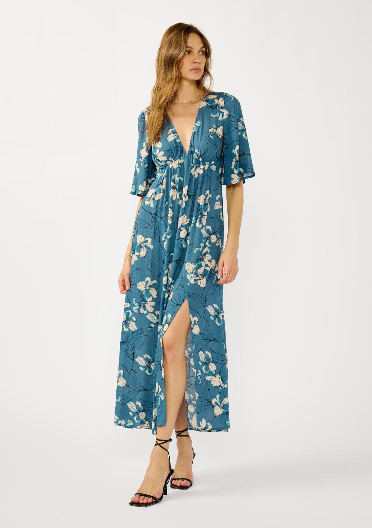 [Color: Teal/Blue] A front facing image of a blonde model wearing a teal floral print empire waist maxi dress with a deep v-neckline, short flutter sleeves, and a chic side slit. An effortless casual fall dress for special occasions.