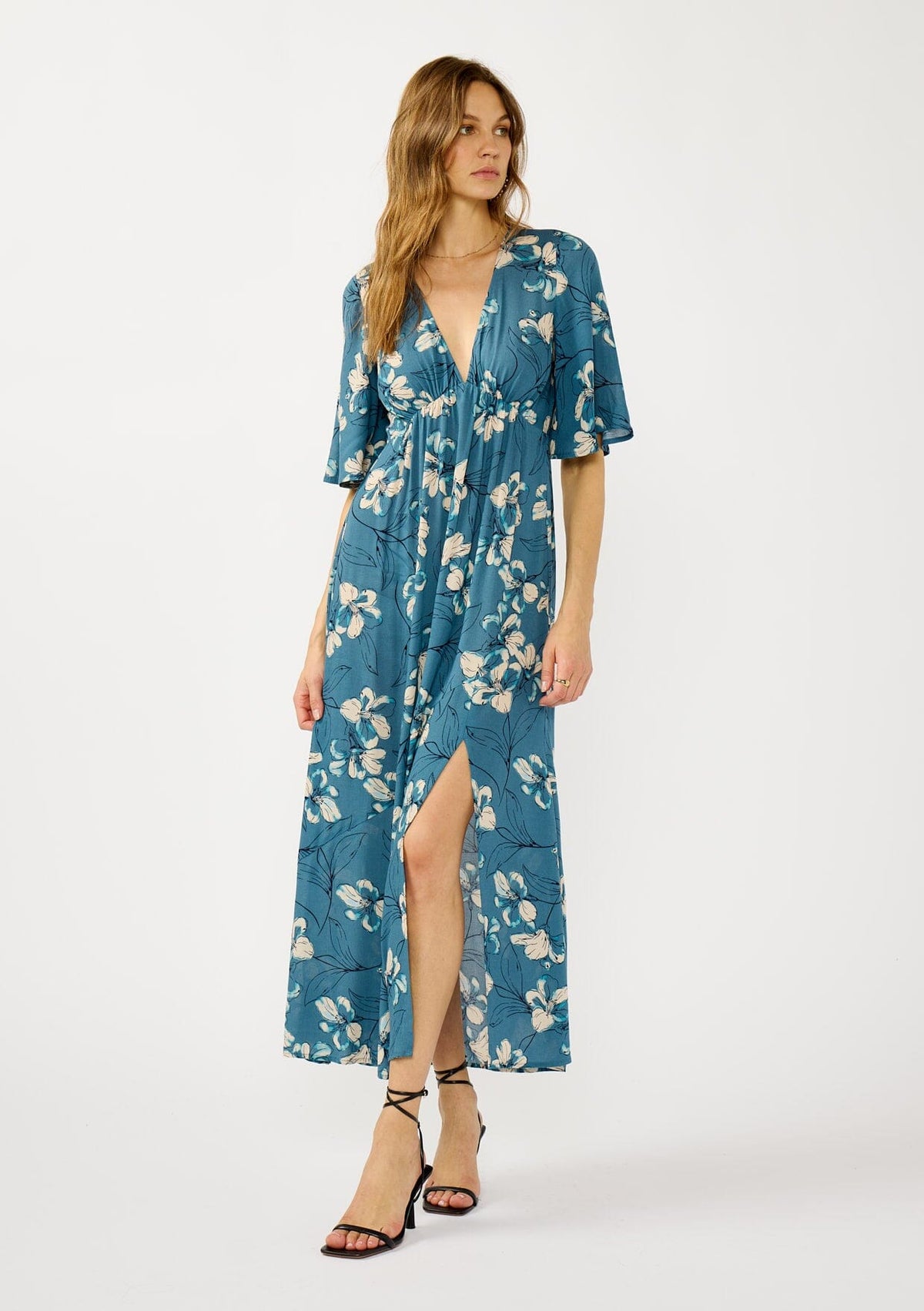[Color: Teal/Blue] A front facing image of a blonde model wearing a teal floral print empire waist maxi dress with a deep v-neckline, short flutter sleeves, and a chic side slit. An effortless casual fall dress for special occasions.