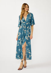 [Color: Teal/Blue] A front facing image of a blonde model wearing a teal floral print empire waist maxi dress with a deep v-neckline, short flutter sleeves, and a chic side slit. An effortless casual fall dress for special occasions.
