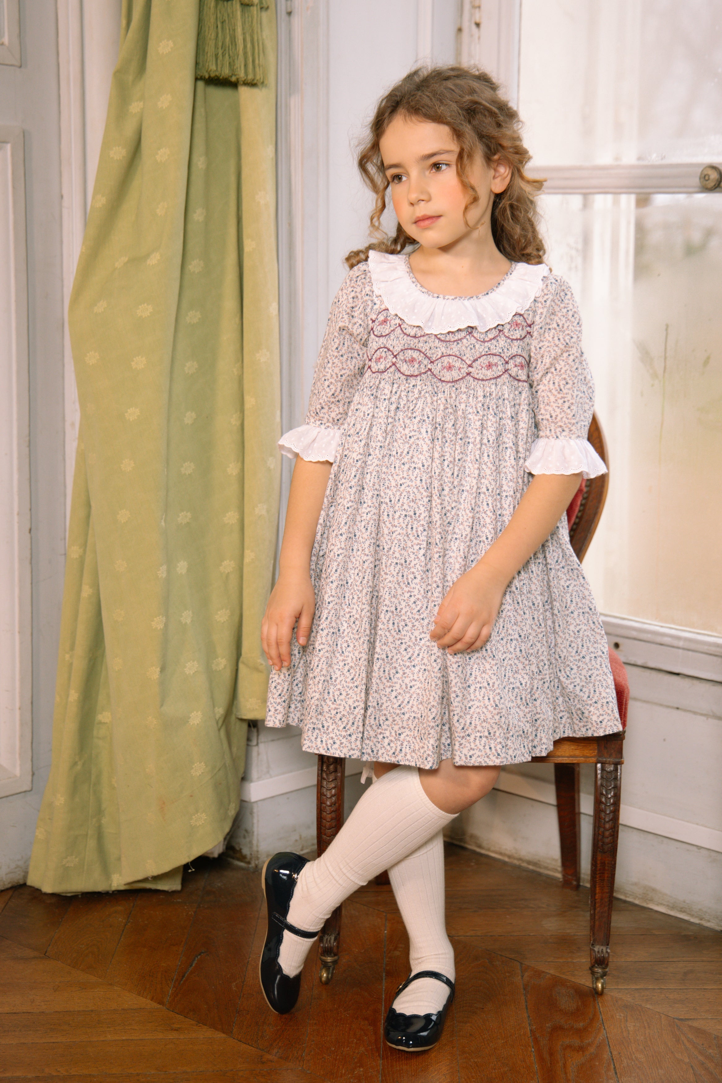 Girl in long-sleeve floral smocked dress with scalloped lace collar and lace-trim cuffs, posing in a Parisian room.