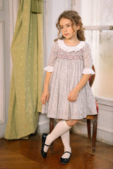 Girl in long-sleeve floral smocked dress with scalloped lace collar and lace-trim cuffs, posing in a Parisian room.