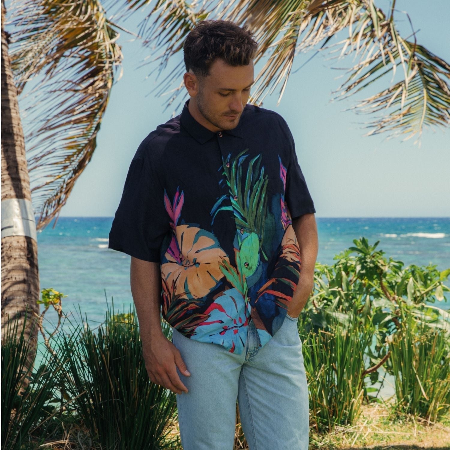 Man wearing a colorful tropical shirt standing in front of palm trees and the ocean.