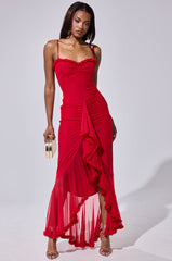A woman in the TATI RUFFLE MAXI DRESS IN RED—fitted with thin straps, ruching, ruffle trim, and a sheer asymmetrical skirt—holds a gold clutch. She wears strappy heels and has straight, shoulder-length hair.