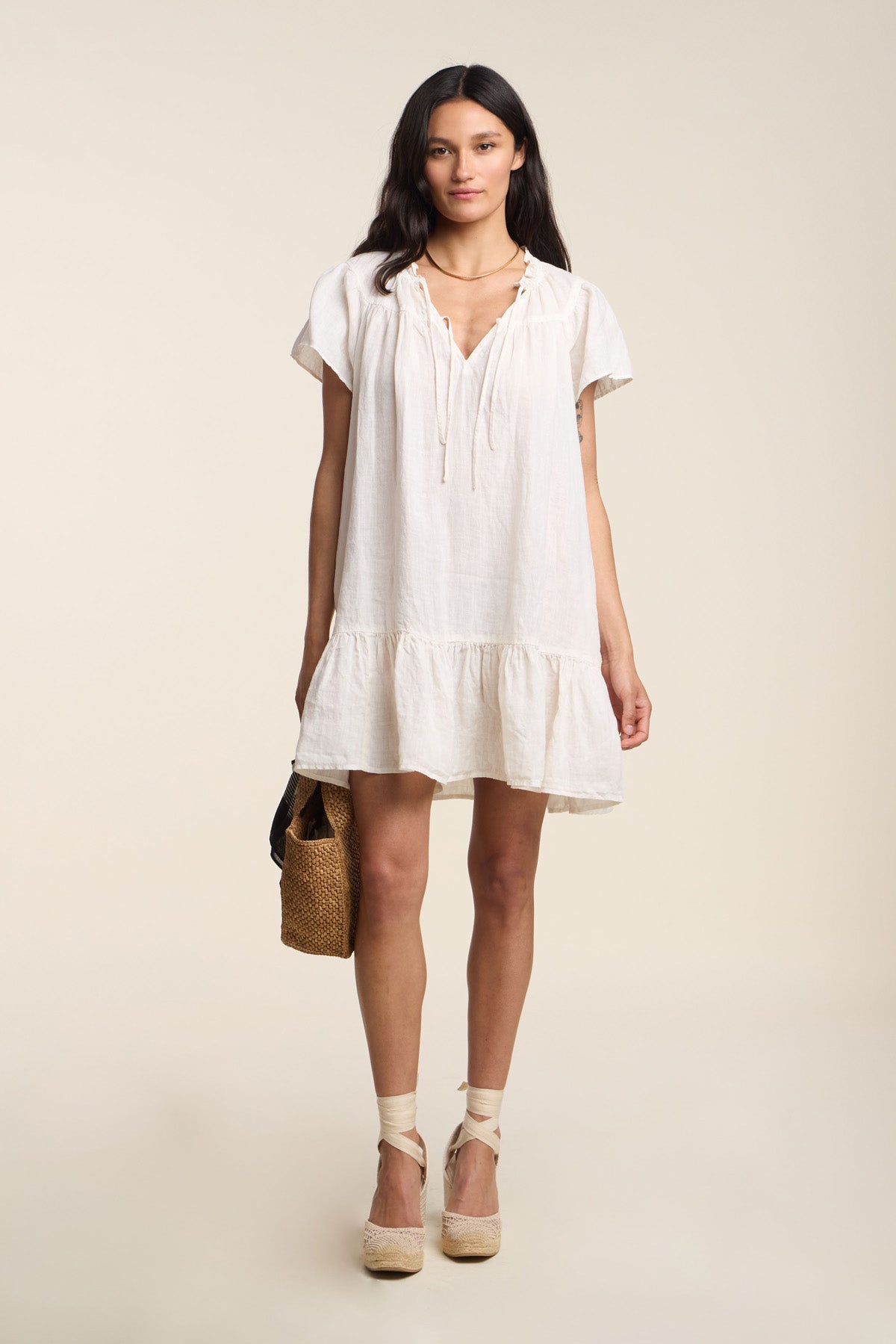 A woman faces forward wearing the LIVVY LINEN DRESS by Velvet by Graham & Spencer with short sleeves and a loose fit, paired with wedge sandals and holding a woven handbag.
