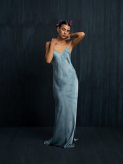 A woman in a flowing Lily Silk Dress - Cosmic Indigo Tiedye by Rimmba | Sustainable Clothing stands against a dark background, eyes closed, with one arm behind her head and small pink flowers in her hair.