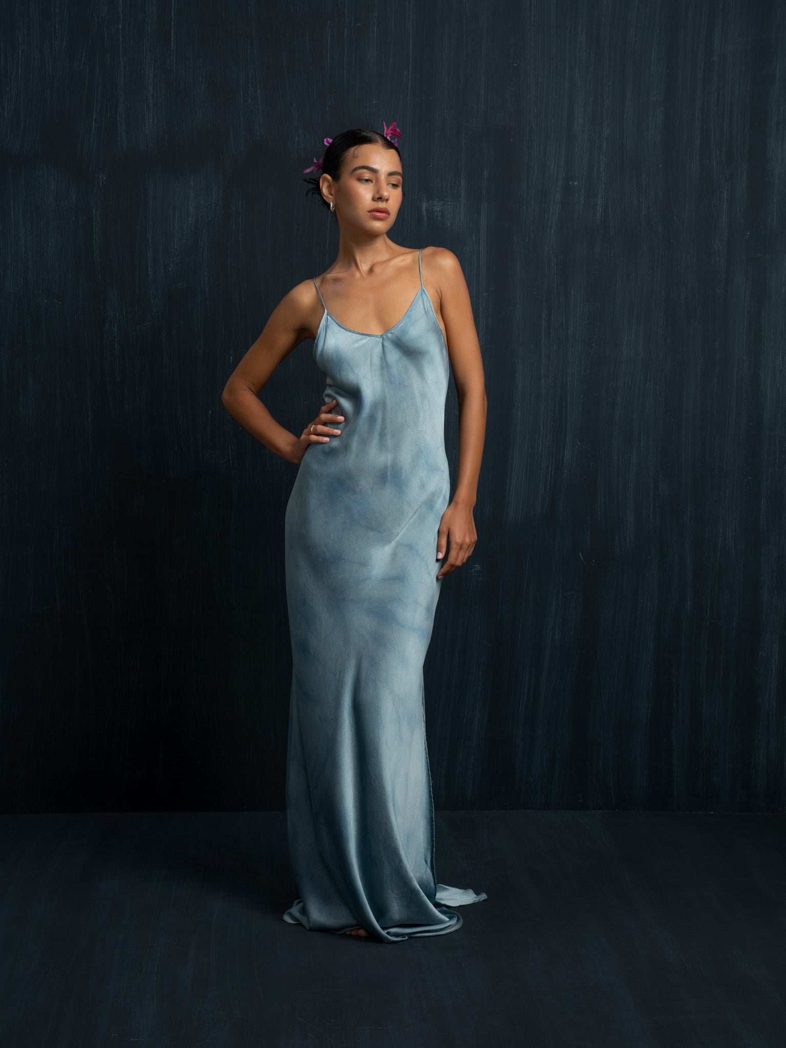 A woman poses against a dark backdrop in the Lily Silk Dress - Cosmic Indigo Tiedye by Rimmba | Sustainable Clothing, exuding effortless elegance with floral accents in her hair and a hand on her hip.