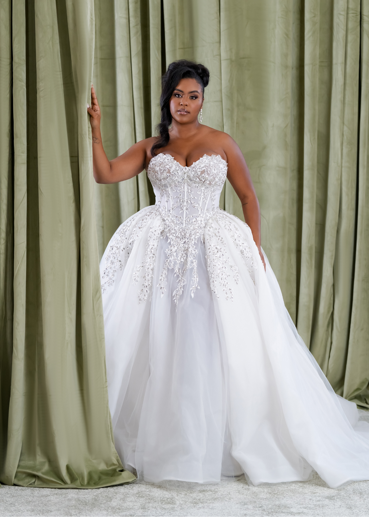Plus size Black bride wearing a strapless ballgown wedding dress with a sheer corset bodice, exposed boning, crystal appliqué, and full organza skirt, designed by Black bridal brand Pantora.