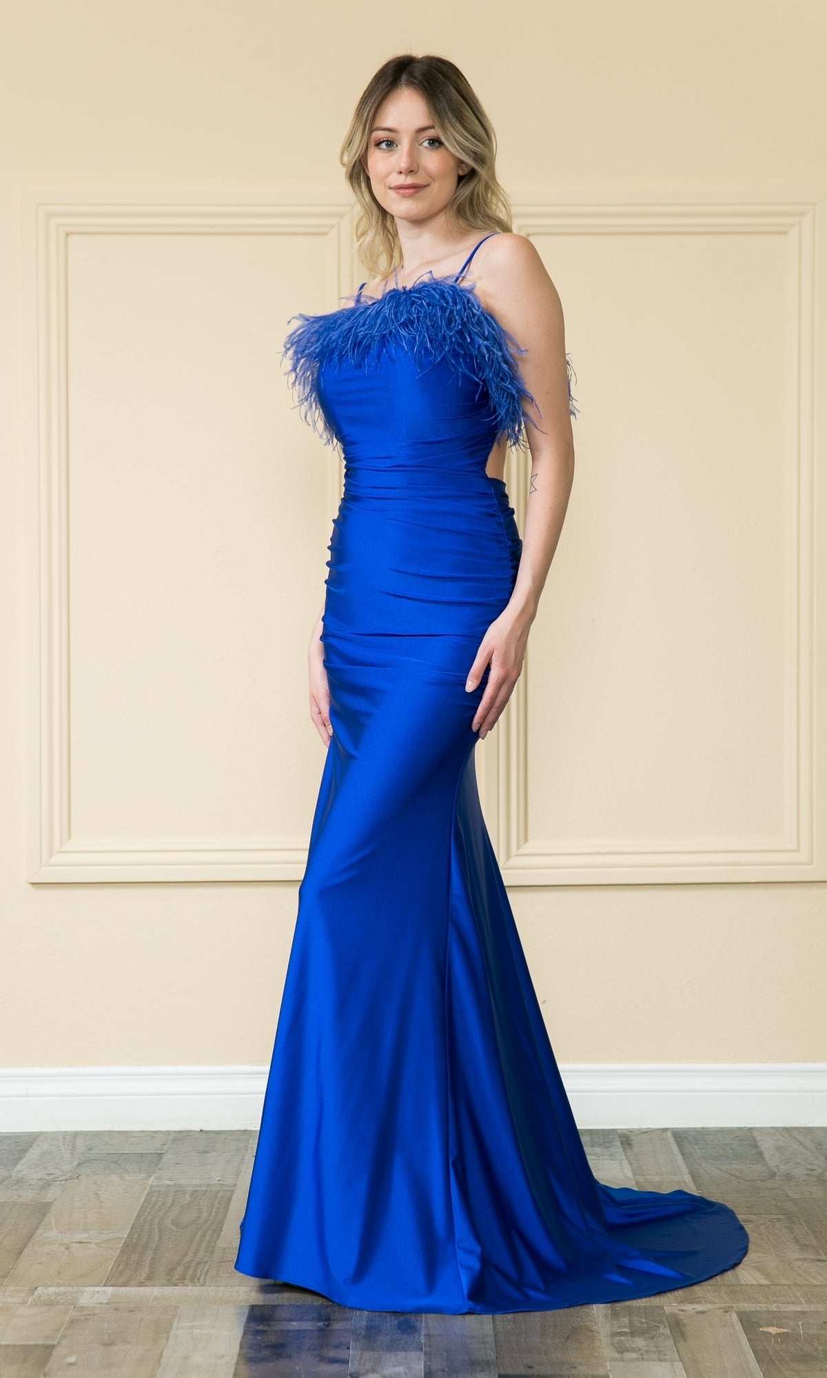 A woman in the affordable Open-Back Long Feathered Prom Dress 9070 in royal stands on a wooden floor indoors against a beige wall with decorative molding. The fitted, floor-length gown features feathered trim for an elegant formal look.