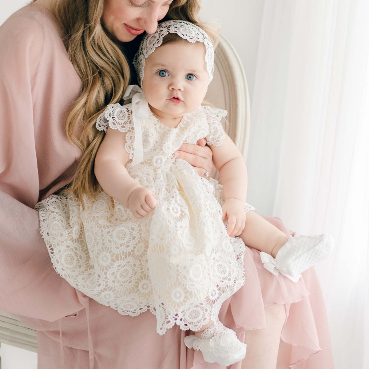 A baby girl dressed in the Poppy Blessing Dress, featuring detailed floral lace and a matching lace headband, seated on her mother’s lap, with soft lighting accentuating the dress's elegance.