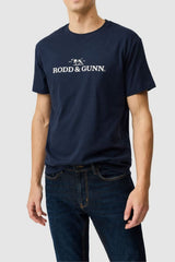 A man wears the Rodd & Gunn The Logo Tee 2.0 in navy blue, featuring white text and a small logo on 100% cotton, paired with dark blue jeans, standing against a plain light gray background.