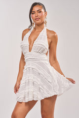 A woman models the CARMEN RUFFLE MINI DRESS, a white halter-neck mini dress with ruffles, against a plain light background. She accessorizes with long earrings and has her hair in a side ponytail.
